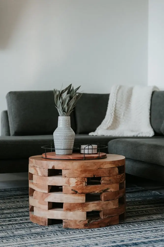 A round stacked-wood coffee table holds a tray, a patterned grey ceramic vase with a leafy plant and a coaster set, in front of a grey sofa with a white knit throw.