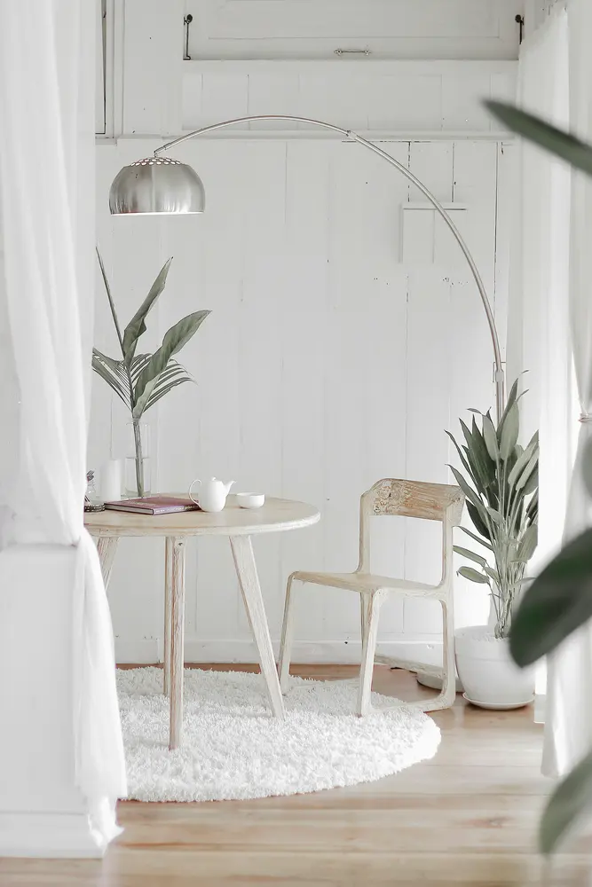 A minimalist white interior nook with a round wooden table, chair, arc floor lamp, and potted plant.