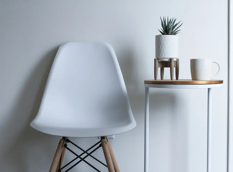 A white chair next to a slim side table holding a plant and a mug