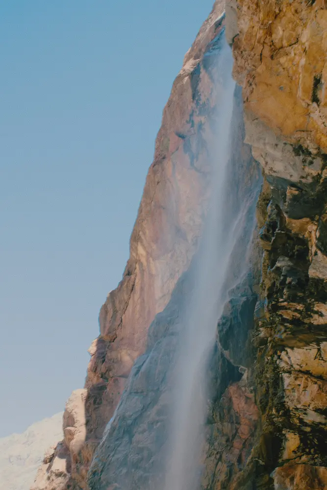 A waterfall falling down a sunlit rock face below a snow-capped peak